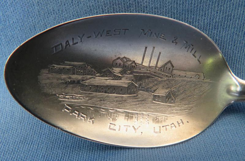Daly West Souvenir Spoon Bowl Marking.JPG - SOUVENIR MINING SPOON DALY WEST MINE & MILL PARK CITY UT - Sterling silver souvenir mining spoon, bowl engraved DALY-WEST MINE & MILL PARK CITY, UTAH with a mining scene of mine and mill, length 6 in., weight 20 gms., handle features Lily of the Valley pattern (1885), monogrammed on back with script Q, Sterling mark and Whiting hallmark (The Park City District was one of Utah’s major mining areas along with the West Mountain (Bingham) and Tintic Districts.  The town of Park City is located 30 miles southeast of Salt Lake City in the Wasatch Mountains.  The town was originally called Parley’s Park when silver was first discovered in 1868.  With a population of 164 in 1870, the town continued to grow and the name was changed to Park City in 1872.  Several successful silver and lead mines were started including the Daly West Mine.  It is located in Summit county at an elevation of 8,376 feet and situated 2 miles south-southwest of Park City, near the head of Empire Canyon.  During the development of the Daly mine it was determined that the great Ontario lode extended westward beyond the limits of the Daly property.  This led to active prospecting and locating during the mid-1880s around the head of Empire Canyon.  The original holdings of the Daly West property embraced 40 patented lode claims, an undivided half being owned by J. B. Haggin, George Hearst, R. Chambers, et al. and the other half by John J. Daly.  The owners began systematic development of their property in the summer of 1891. By the end of 1892, the shaft had reached a depth of 1,255 feet and was connected with the west drift from the Daly mine. In October 1893, Daly incorporated his one-half as the Daly West Mining Co. with $1,500,000 capitalization in $20 shares under the laws of Utah. Soon after, the other half was acquired by the Ivanhoe Mining Co. of California.  Rich ore was found in the summer of 1894 and a period of extensive development and active mining followed. Throughout the following year the output increased significantly and in August 1895 the concentration mill was constructed.  Early in 1899 the Daly West and Ivanhoe companies were consolidated under the name Daly West Mining Co.  The total product of the Daly West from 1893 to 1906 sold for $11.2 million.  Park City was incorporated in 1884 and by 1896, when Utah became a state, the town had grown to 7,000 people.  Most of the mines closed in 1949, but some briefly opened for a bit in 1952. Though the mines opened again for a bit in the 1980s, it was clear that mining as the main stay of the town's economy was ending.  The idea of having a ski resort in Park City was born in 1963. Treasure Mountain Ski Resort gained some popularity in 1966 when Sports Illustrated gave it high marks in the quality of its runs. Soon, other ski resort outfits established themselves and Park City was reborn economically.)